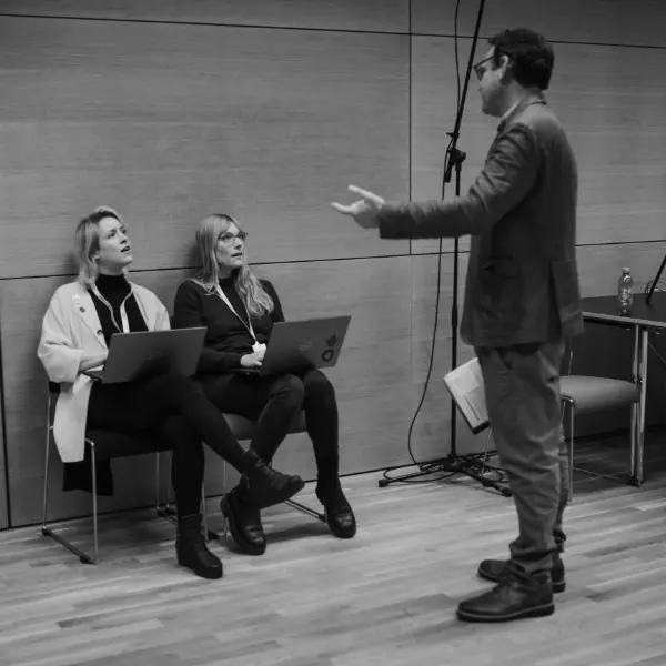 Black-and-white image: man speaks to two seated people with laptops in a room.