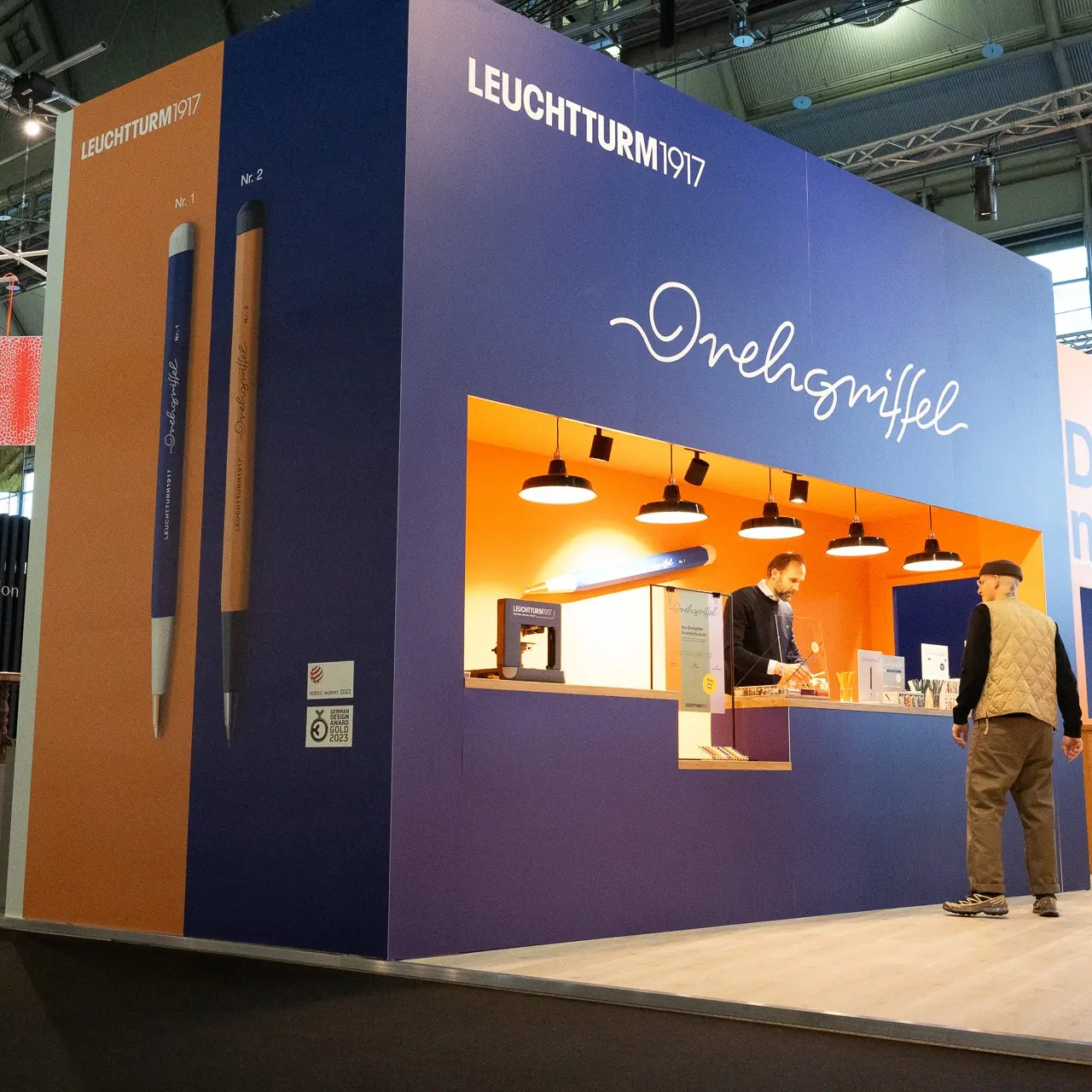 Messestand von LEUCHTTURM1917 auf der Ambiente 2024 mit großem Drehgriffel-Branding, beleuchtetem Counter und zwei Personen im Gespräch vor der Theke.