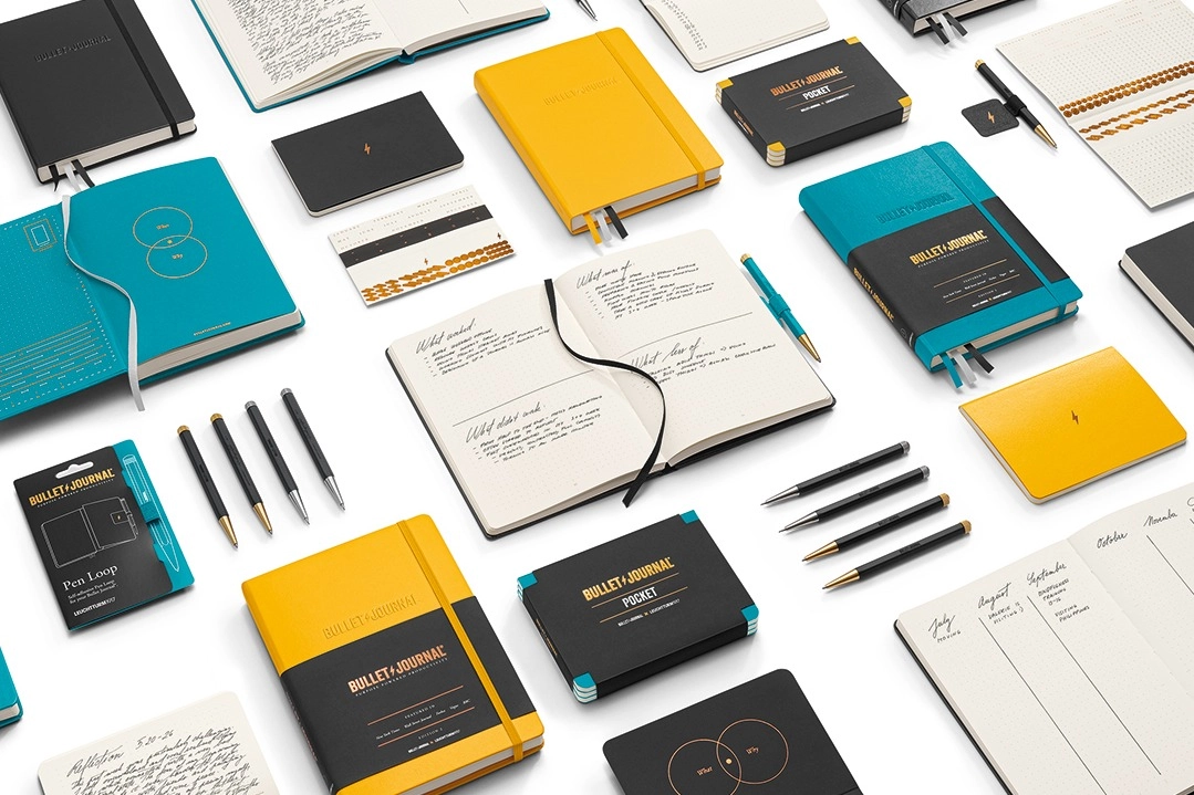 Verschiedene Produkte des Bullet Journal Sortiments in Schwarz, Gelb und Türkis, darunter Notizbücher, Pocket-Formate, offene Beispielseiten, Stiftsets, Pen Loop und Zubehör, arrangiert auf weißem Hintergrund. Bullet Journal notebooks in turquoise, yellow and black with open pages, Drehgriffel and accessories, zoomed in.