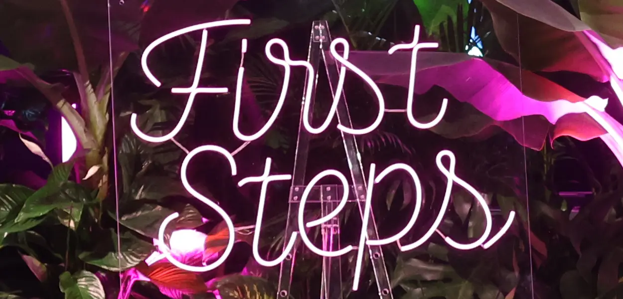 First Steps Award. Neon sign with text “First Steps” in front of plant wall.