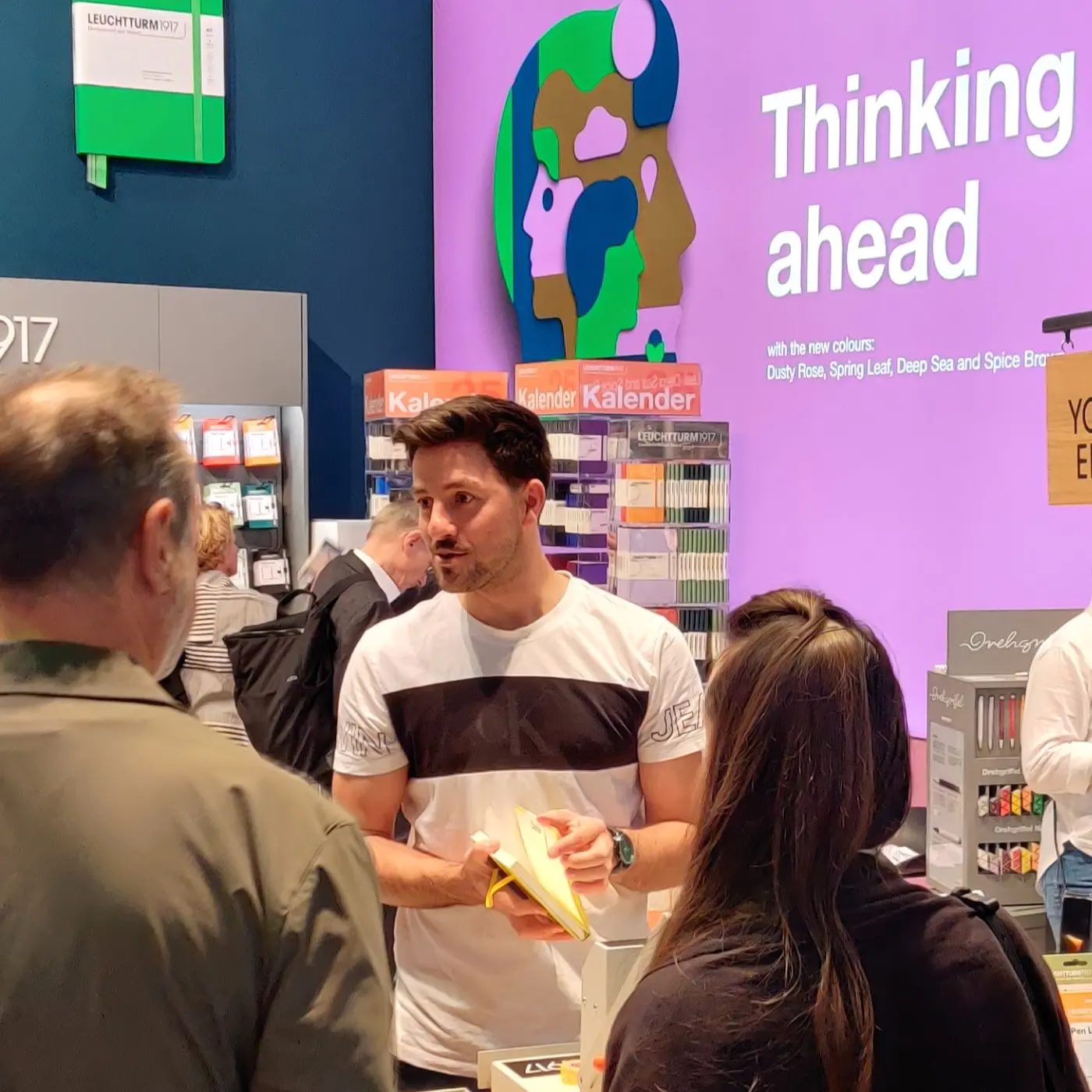 Man talking to visitors at a stand, holding notebook, wall text “Thinking ahead”.