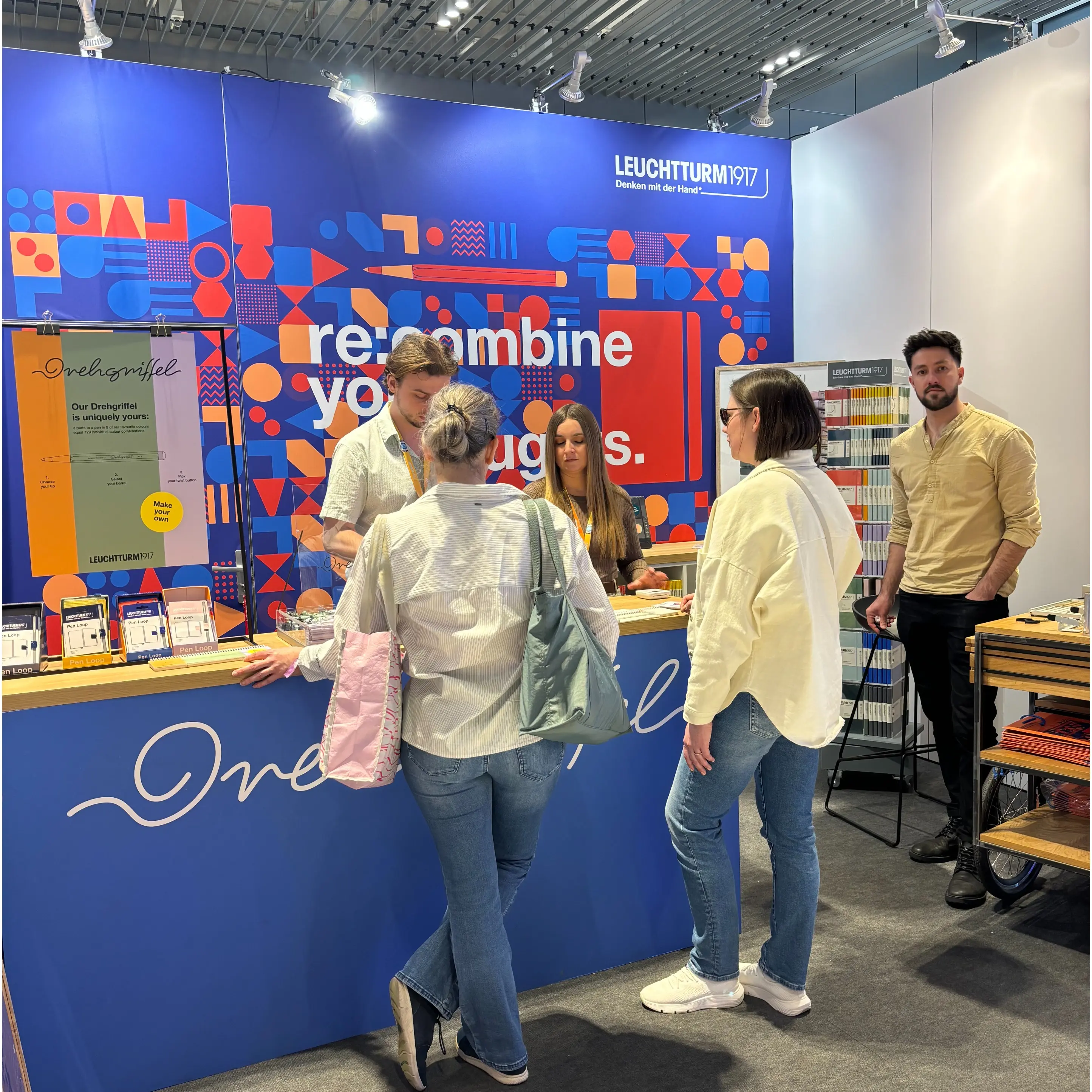 Leuchtturm1917 Messestand Founder Summit 2024. Several people at a trade fair stand with counter, product shelves and wall graphic.