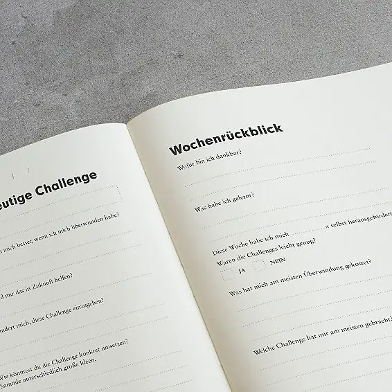 Geöffnete Seiten des Change Journals mit Vorlagen für den Wochenrückblick und eine tägliche Challenge, aufgenommen auf grauem Hintergrund.