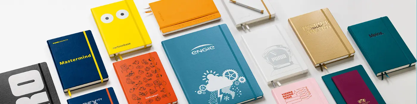 LEUCHTTURM1917 Notizbücher personalisieren, Header. Colorful notebooks with logos, illustrations and embossing, laid out flat in a row.