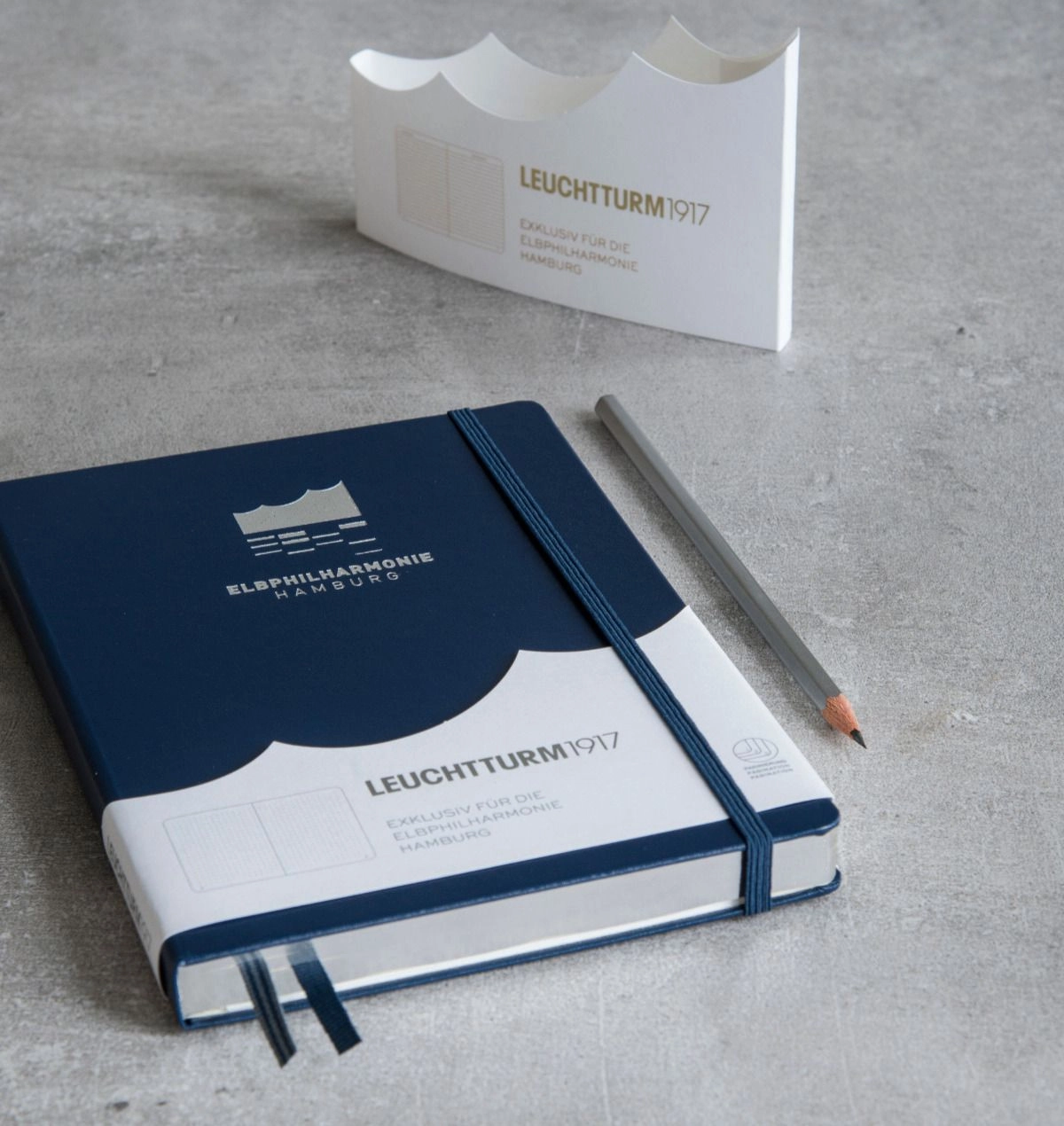 Dark blue notebook with embossing “Elbphilharmonie Hamburg”, elastic band and building-shaped banderole.