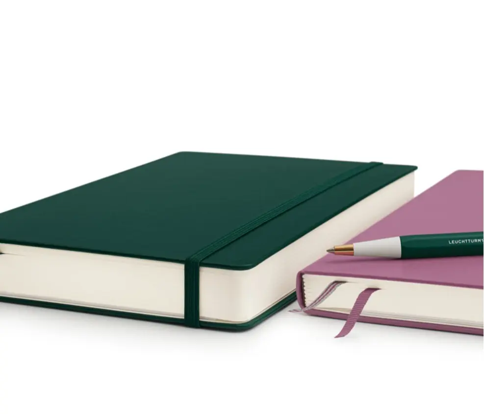 Das Leuchtturm1917 Notizbuch mit 411 Seiten und Platz für noch mehr Ideen. Green and pink notebooks with elastic band and bookmark, next to a green Drehgriffel Nr. 1.