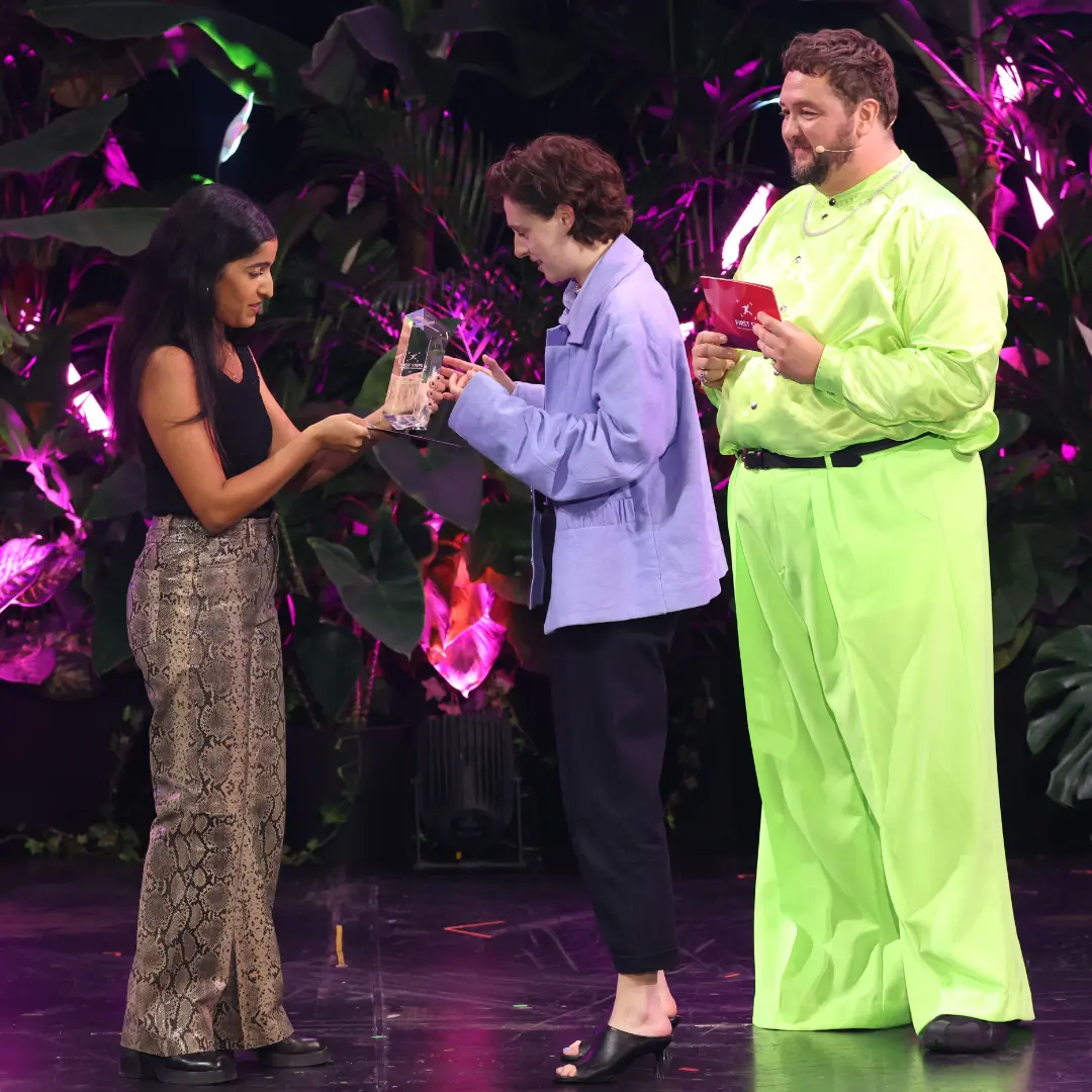 Three people on stage at award ceremony, trophy being handed over.