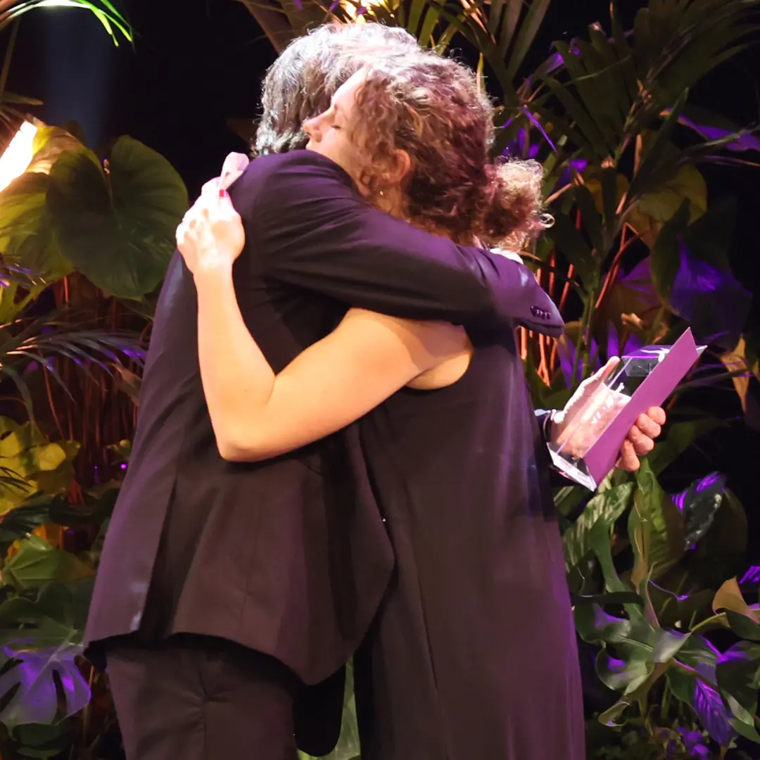 Two people on stage hugging, one holding an award in their hand.