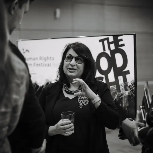 Black-and-white: woman holding a glass talks to people in front of a display wall.