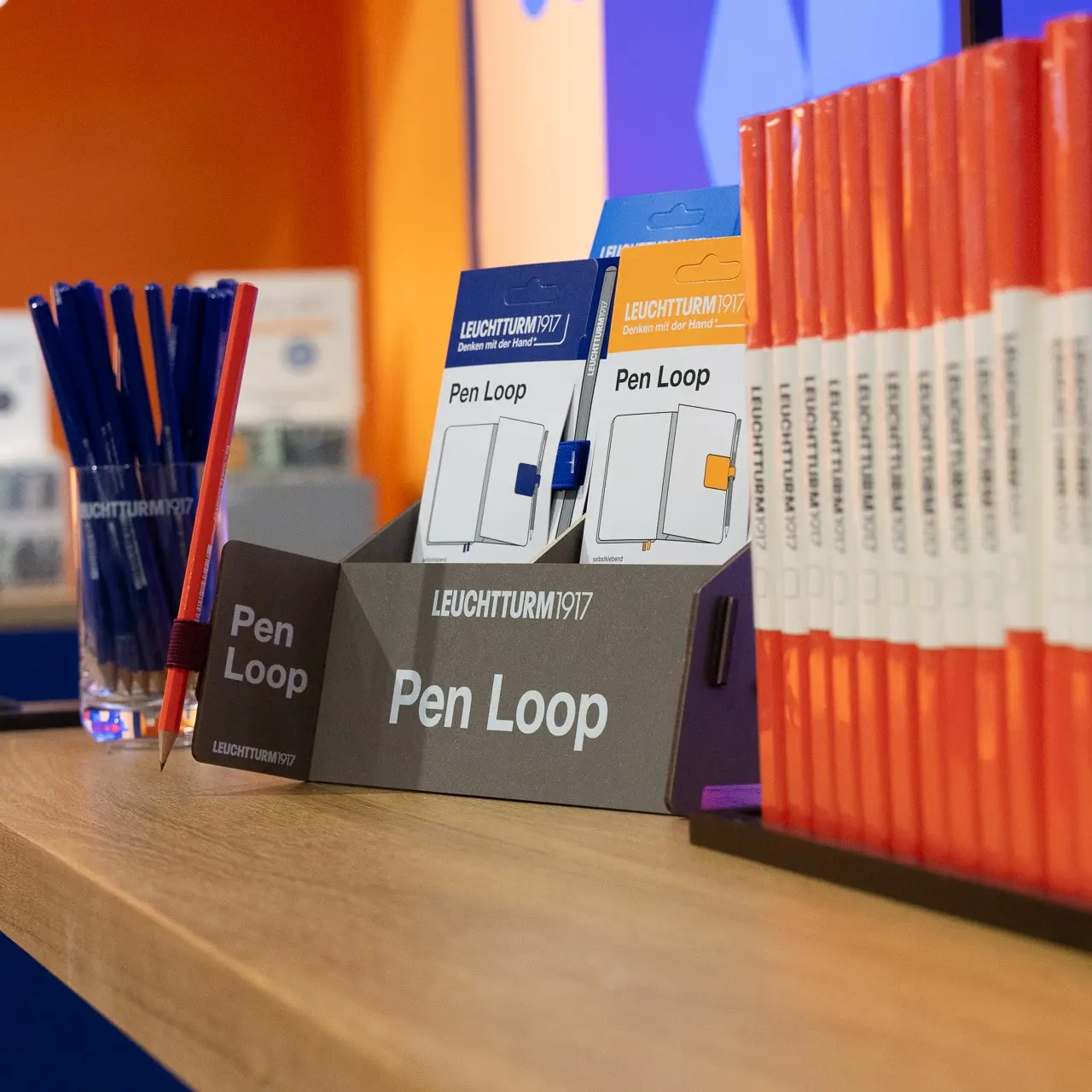 Nahaufnahme eines Pen-Loop-Displays von LEUCHTTURM1917 auf der Ambiente 2024, mit verschiedenen Pen-Loop-Verpackungen und Stiften in Blau und Orange auf einem Holztresen.