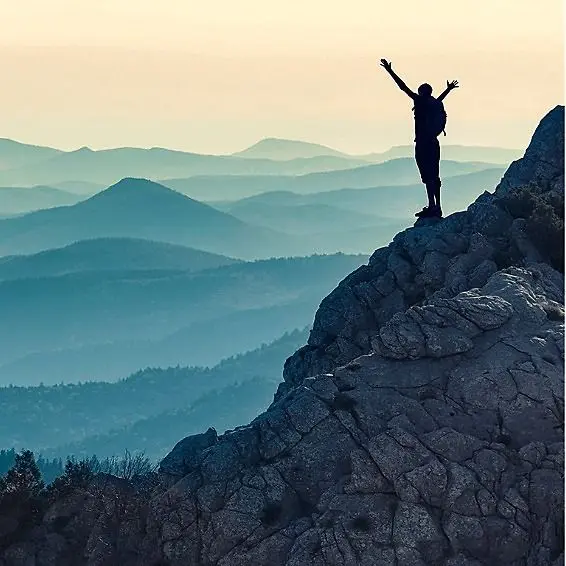 Person steht jubelnd auf einem Felsen mit Blick über mehrere Bergketten bei Sonnenaufgang.