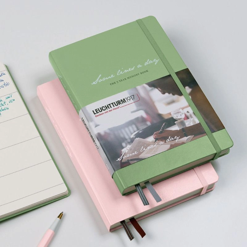 Two notebooks in green and pink with embossing, elastic band and banderole, book and Drehgriffel beside.