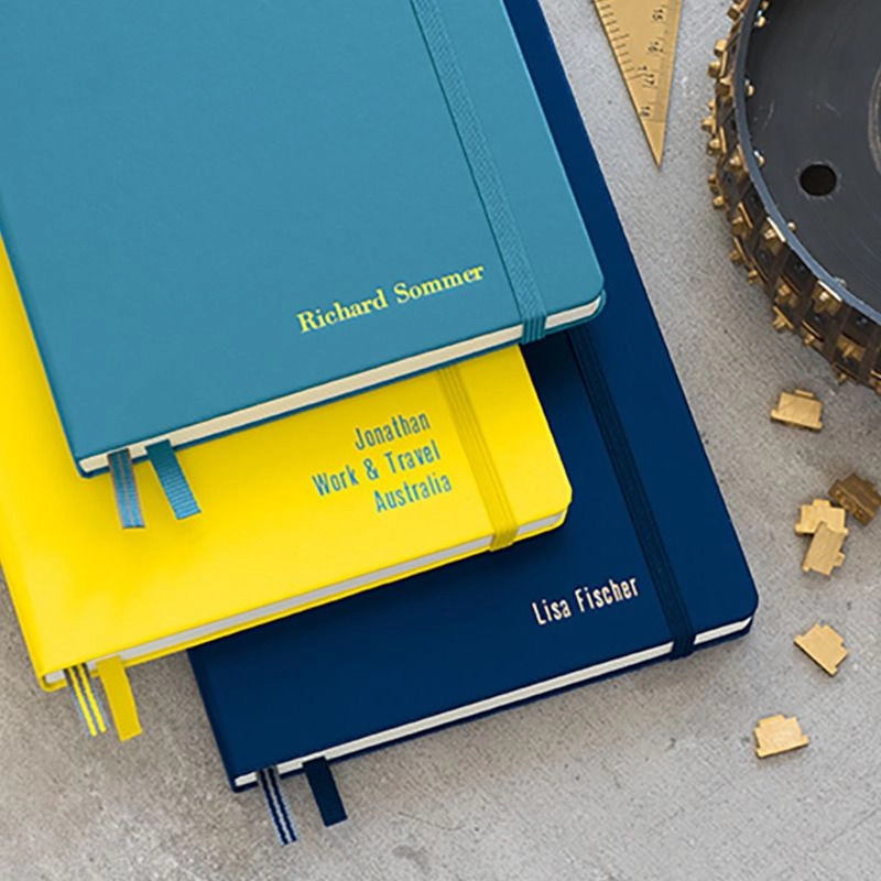 Notizbuch_Prgung Three notebooks in turquoise, yellow and dark blue with elastic band and embossings, metal parts beside.