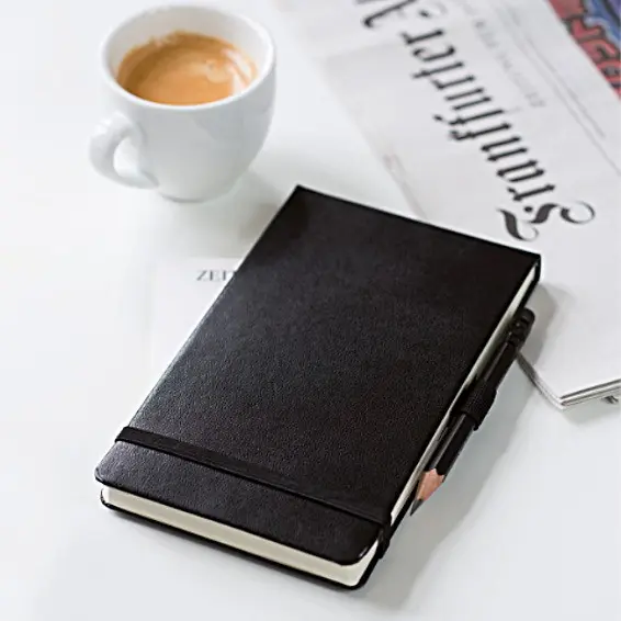 Der praktische Leuchtturm1917 Notizblock, perfekt für unterwegs. Black notebook with attached pencil, next to a cup of coffee and a newspaper.