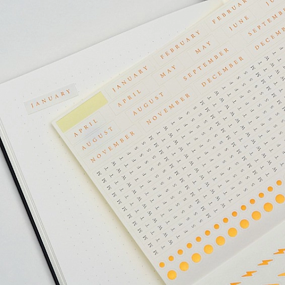 Bullet Journal – Stickerbogen mit Monats- und Symbolstickern Dotted notebook page with monthly sticker sheet January–December, orange dots and lightning symbols.