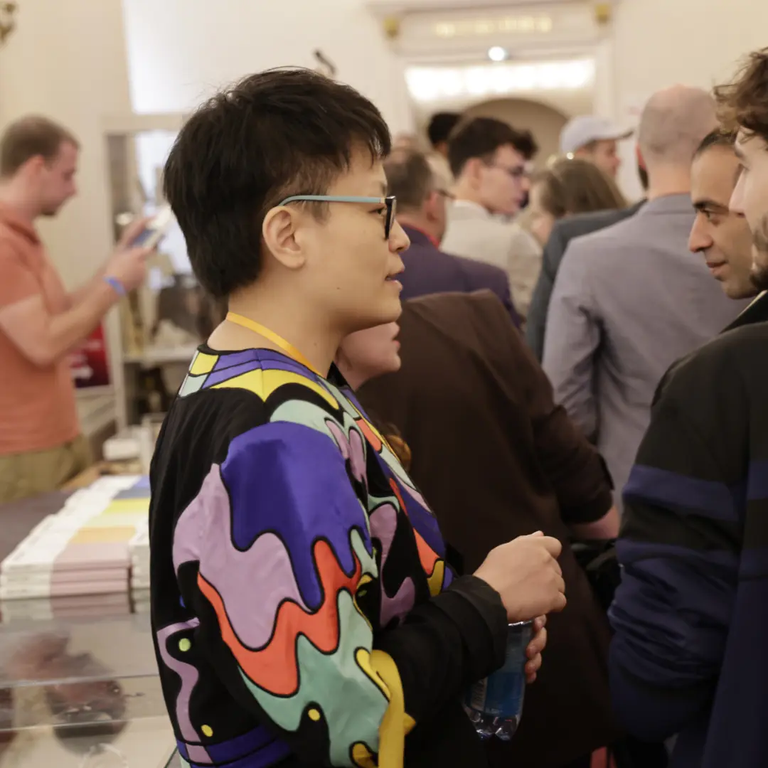 Person talking at the trade fair stand, colorful top and crowd in the background.