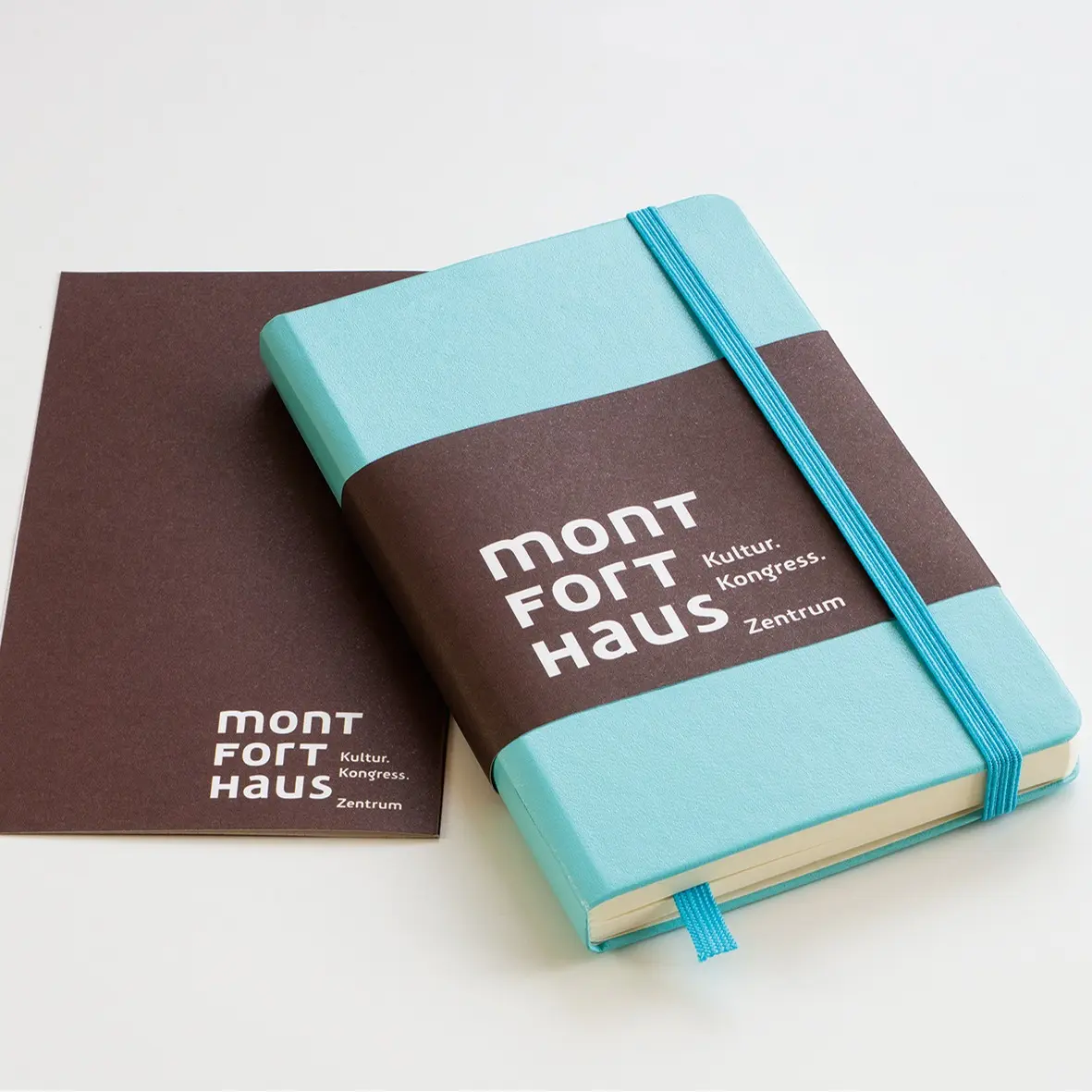 Turquoise notebook with brown band “montfort haus”, elastic band and brown booklet in background.