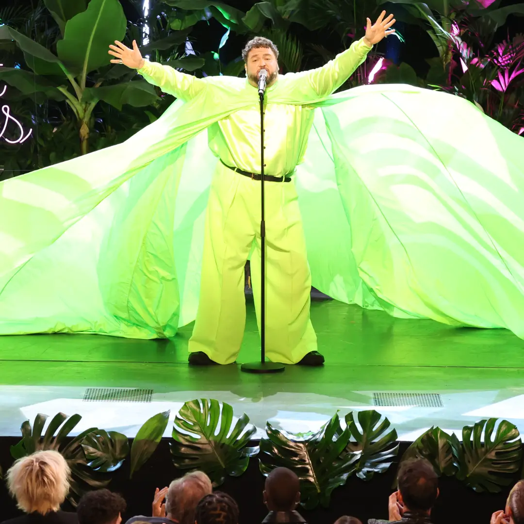 Man on stage with microphone, arms outstretched and green fabric cape.