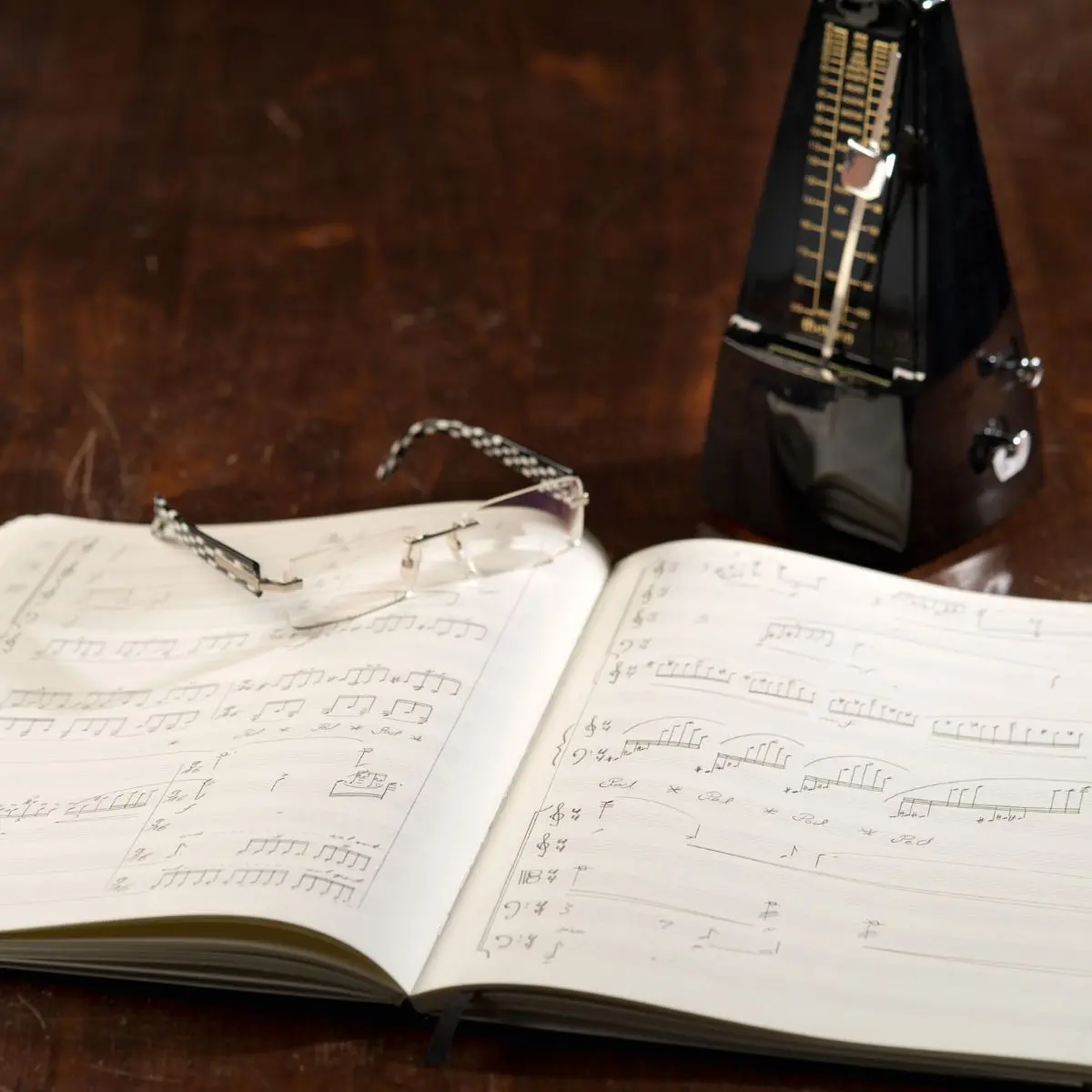 leuchtturm1917-notenbuch Aufgeschlagenes Buch mit handschriftlichen Musiknoten, Brille darauf, daneben Metronom.