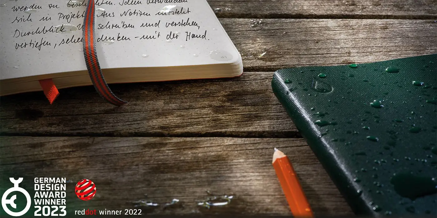 leuchtturm1917_outlines_hero Open and closed notebook with water droplets, pencil and two design awards, on a wooden surface.
