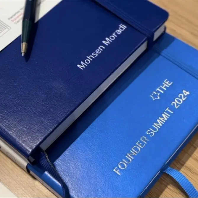 Detailaufnahme personalisierte Leuchtturm1917 Notizbücher auf dem Founder Summit 2024. Two blue LEUCHTTURM1917 notebooks embossed “Mohsen Moradi” and “FOUNDER SUMMIT 2024”.