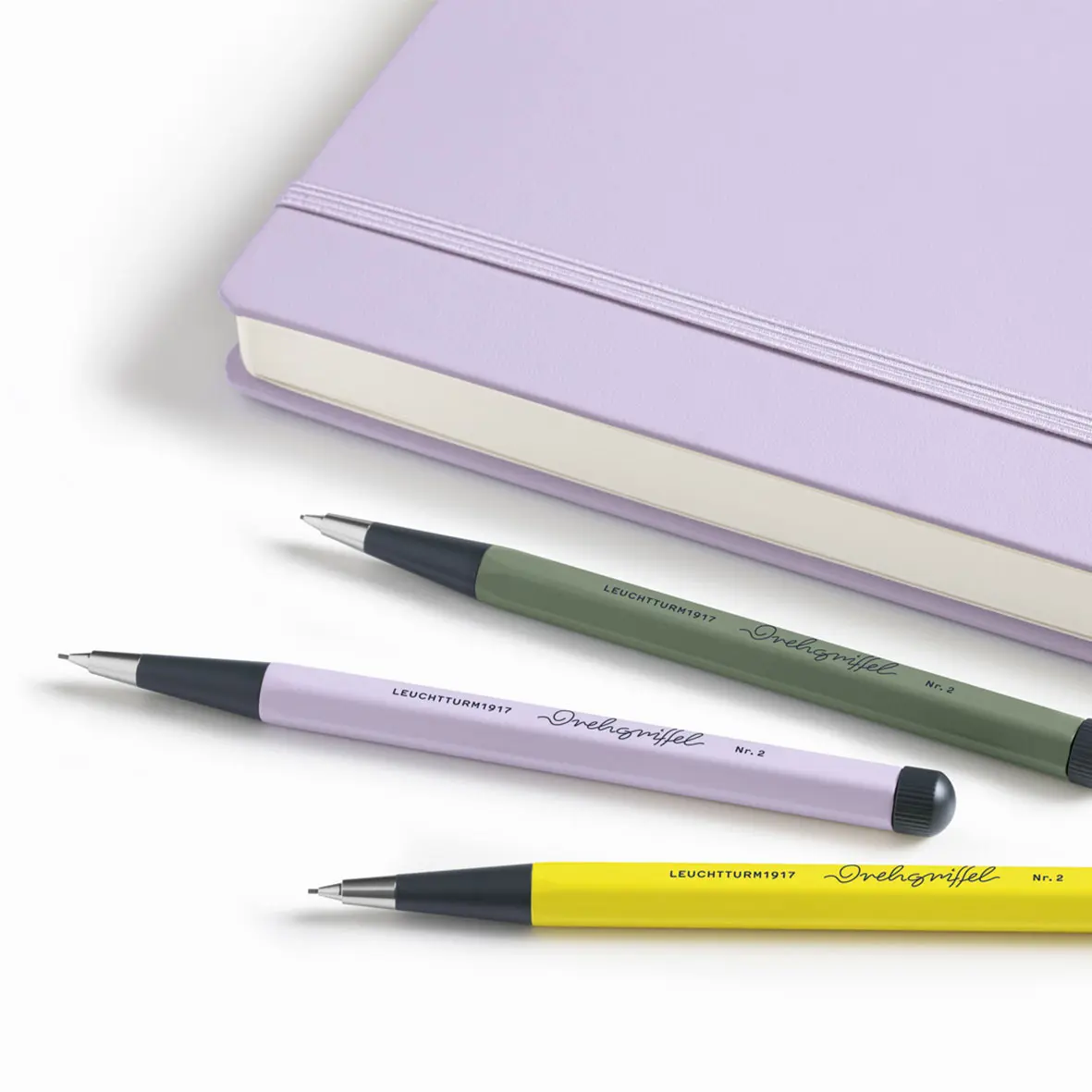 Drehgriffel Nr. 2 – Lilac, Olive & Zitrone mit Notizbuch in Lilac. Drei metallene Bleistift in Grün, Flieder und Gelb vor fliederfarbenem Notizbuch.
