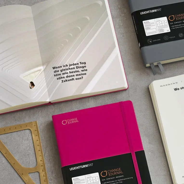 Mehrere Change Journal Notizbücher in Pink und Grau sowie eine geöffnete Doppelseite mit inspirierendem Zitat, arrangiert auf grauem Untergrund.