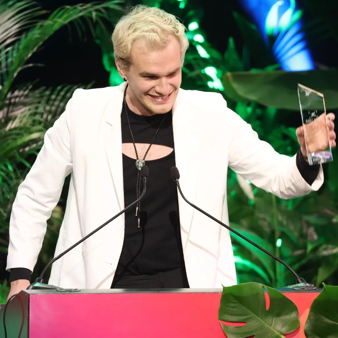 Man on stage holding a trophy at a lectern, plants in the background.