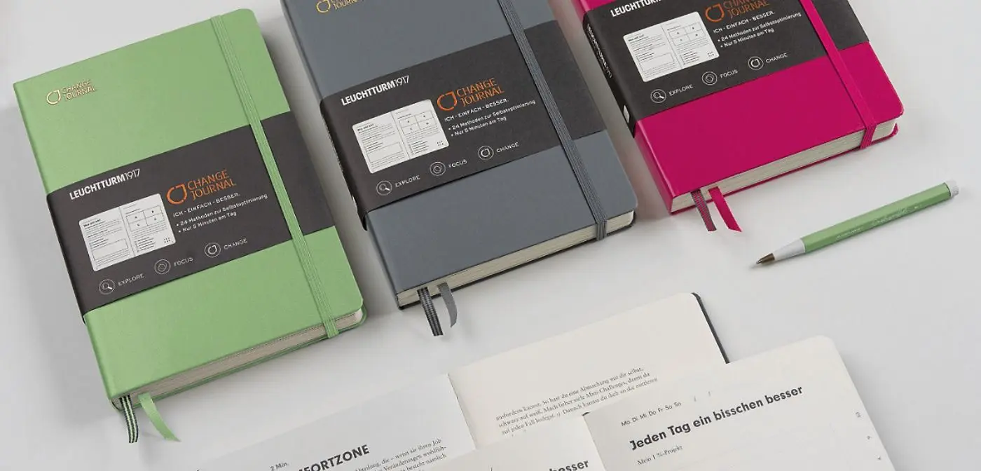 Drei Change Journal Notizbücher in Grün, Grau und Pink mit schwarzer Banderole, dazu ein passender Stift und offene Beispielseiten auf hellem Hintergrund.