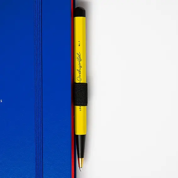 Bauhaus Edition - Drehgriffel No. 1 on notebook with Pen Loop. Blue notebook with red page edges, yellow-black ballpoint pen with gold-coloured tip attached.